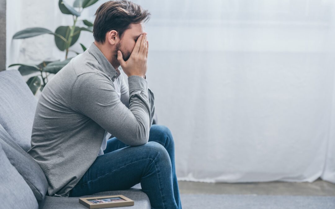 man sitting on grey couch with photo in frame and crying at home, grieving disorder concept man sitting on grey couch with photo in frame and crying at home, grieving disorder concept